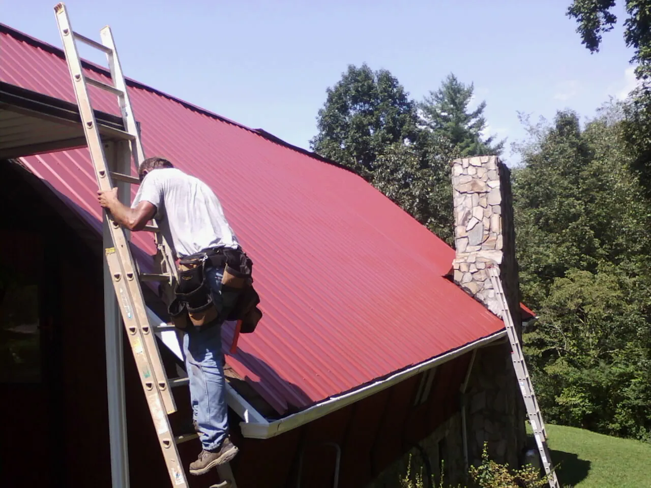 Roofer on a ladder beside a red metal roof and stone chimney — Soffit & Fascia Repair by Roofing Brothers in Marston